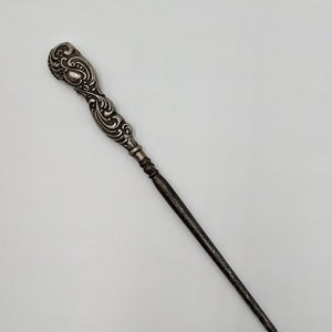May include: An ornate, decorative, metal bodkin with a long, thin, pointed end, finished in silver.