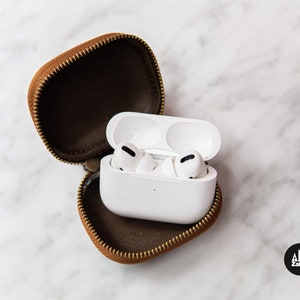 May include: White wireless earbuds in a brown leather case with a zipper. The earbuds are in their charging case.