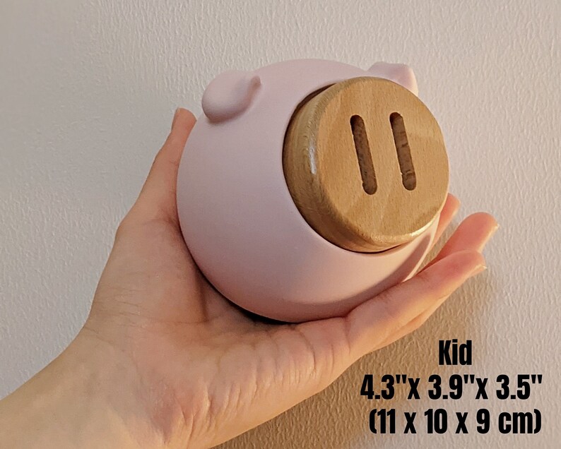 May include: A pink ceramic piggy bank with a wooden coin slot. The piggy bank is 4.3 inches by 3.9 inches by 3.5 inches.
