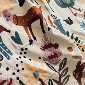 Puede incluir: Tela color crema con una escena de bosque caprichosa. El dise&ntilde;o presenta ciervos, p&aacute;jaros, conejos y elementos florales en tonos marrones, azules y naranjas. La tela tiene una apariencia texturizada.