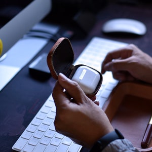 May include: A brown leather case with a zipper closure, open to reveal a white wireless earbud charging case.