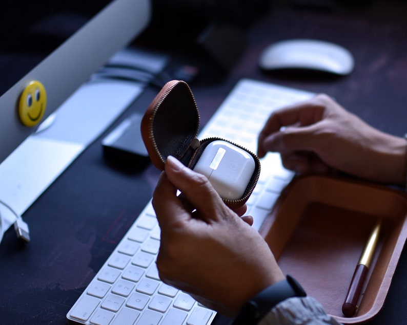 May include: A brown leather case with a zipper closure, open to reveal a white wireless earbud charging case.