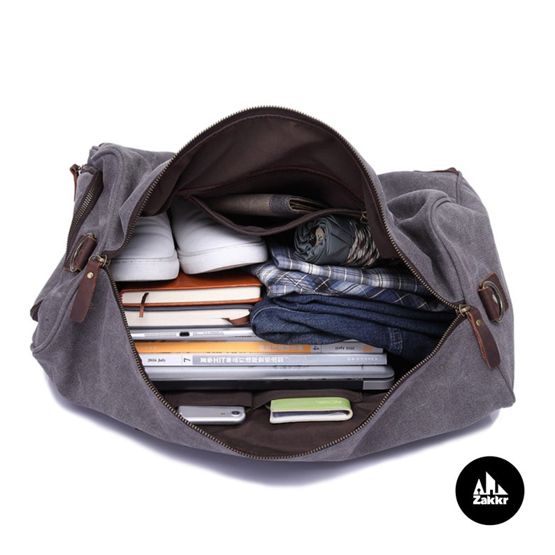 Pu&ograve; includere: Una borsa da viaggio in tela grigia con dettagli in pelle marrone e chiusure a zip. La borsa &egrave; aperta e piena di vari oggetti, tra cui libri, vestiti ed elettronica.