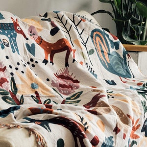 Puede incluir: Una manta crema con un dise&ntilde;o forestal caprichoso. La manta presenta ilustraciones coloridas de ciervos, flores y otros elementos naturales. La manta est&aacute; sobre un sof&aacute;, mostrando sus bordes con flecos y su textura suave.