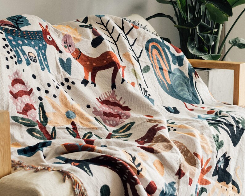 Puede incluir: Una manta crema con un dise&ntilde;o forestal caprichoso. La manta presenta ilustraciones coloridas de ciervos, flores y otros elementos naturales. La manta est&aacute; sobre un sof&aacute;, mostrando sus bordes con flecos y su textura suave.