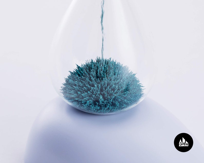 K&ouml;nnte beinhalten: Eine gl&auml;serne Sanduhr mit blauem magnetischem Sand im Inneren. Der Sand ist zu einer Kugelform zusammengeklumpt.