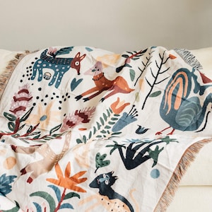 Puede incluir: Una manta crema con un dise&ntilde;o caprichoso de animales y flores. La manta presenta ilustraciones de un zorro, un ciervo y un p&aacute;jaro, junto con varias flores y follaje en tonos de azul, naranja y marr&oacute;n. La manta tiene un borde con flecos.