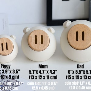 May include: Three white ceramic piggy banks with wooden coin slots. The piggy banks are labeled "Piggy", "Mum", and "Dad" with their dimensions in inches and centimeters.