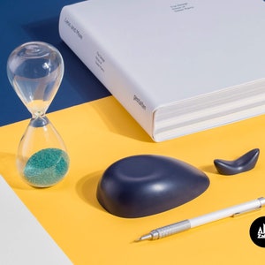 K&ouml;nnte beinhalten: Eine blaue walf&ouml;rmige Schale, eine kleine Walfigur, ein Bleistift und eine Sanduhr mit gr&uuml;nem Sand auf einer gelben Oberfl&auml;che. Der Text "Zakkr" ist in der unteren rechten Ecke des Bildes sichtbar.