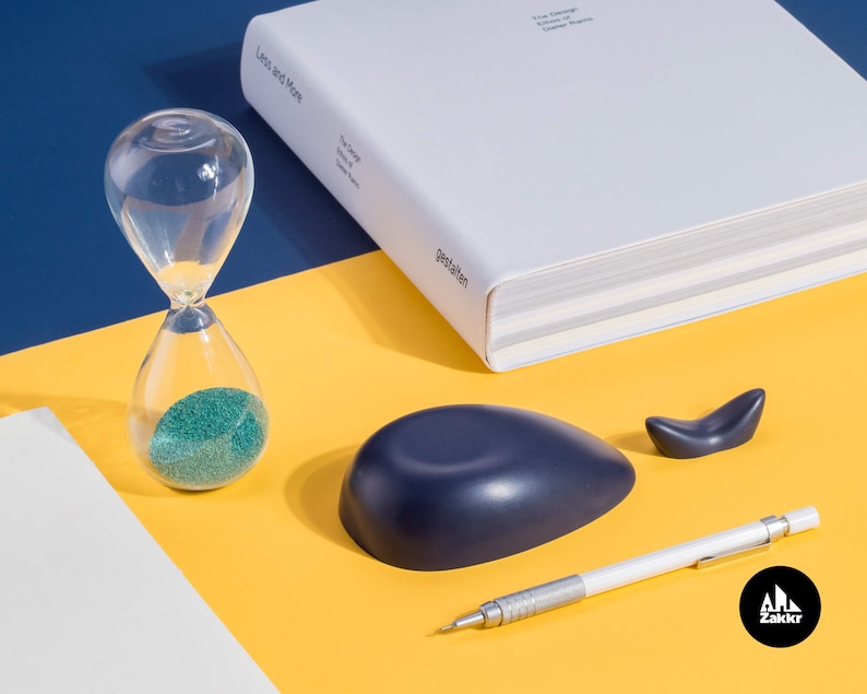 K&ouml;nnte beinhalten: Eine blaue walf&ouml;rmige Schale, eine kleine Walfigur, ein Bleistift und eine Sanduhr mit gr&uuml;nem Sand auf einer gelben Oberfl&auml;che. Der Text "Zakkr" ist in der unteren rechten Ecke des Bildes sichtbar.