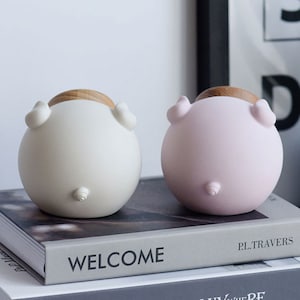 May include: Two ceramic piggy banks, one white and one pink, with wooden tops. The piggy banks are sitting on a book with the word "WELCOME" printed on the cover.