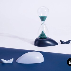 K&ouml;nnte beinhalten: Eine blaue und wei&szlig;e Sanduhr in Form eines Wals, mit gr&uuml;nem Sand im Inneren. Die Sanduhr steht auf einer blauen Oberfl&auml;che mit einer kleineren Walfigur daneben.