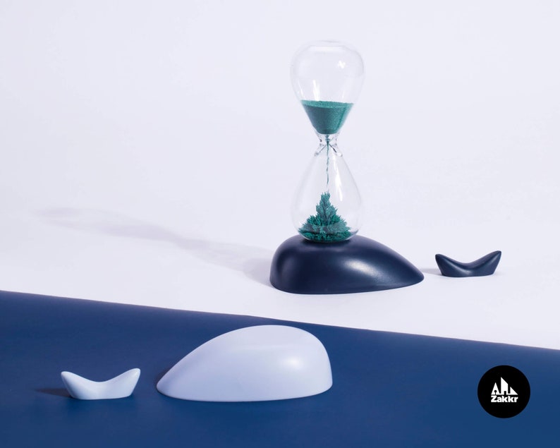 K&ouml;nnte beinhalten: Eine blaue und wei&szlig;e Sanduhr in Form eines Wals, mit gr&uuml;nem Sand im Inneren. Die Sanduhr steht auf einer blauen Oberfl&auml;che mit einer kleineren Walfigur daneben.