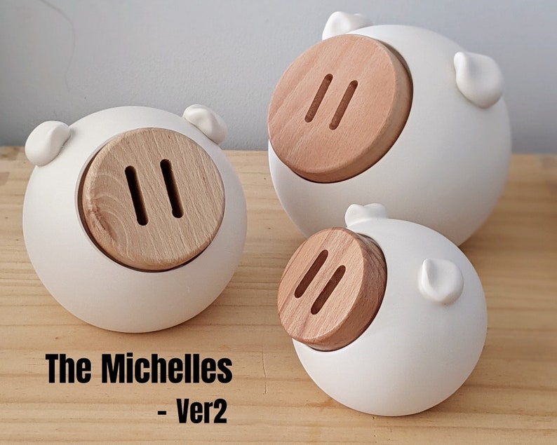 May include: Three white ceramic piggy banks with wooden coin slots. The piggy banks are different sizes and are arranged in a row on a wooden surface. The text "The Michelles - Ver2" is printed below the piggy banks.