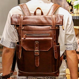 Può includere: Uno zaino in pelle marrone con chiusura a rotolo, spallacci regolabili e una tasca frontale con cerniera. Lo zaino ha un aspetto usurato e vintage.