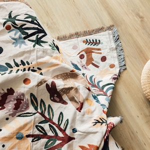 Puede incluir: Una manta tejida de color crema con un dise&ntilde;o caprichoso de animales del bosque y flores. La manta presenta un patr&oacute;n repetido de ciervos marrones, conejos y follaje colorido. Los bordes est&aacute;n acabados con un ribete de flecos. Una cesta tejida est&aacute; en el fondo.