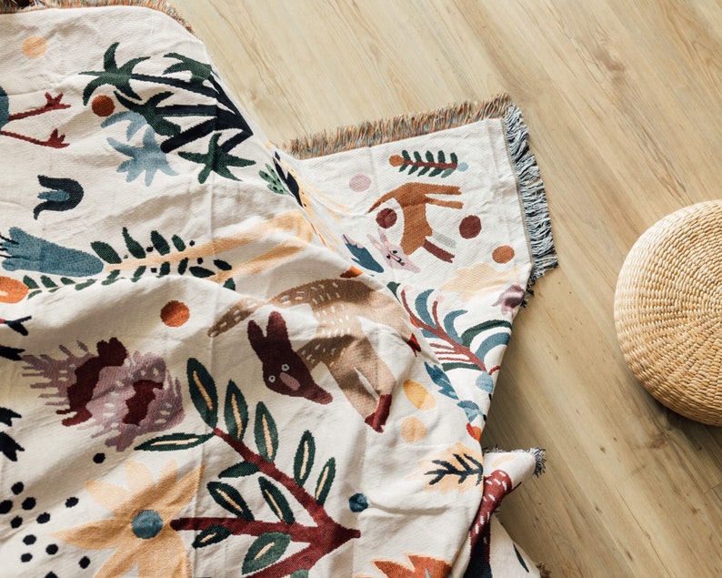 Puede incluir: Una manta tejida de color crema con un dise&ntilde;o caprichoso de animales del bosque y flores. La manta presenta un patr&oacute;n repetido de ciervos marrones, conejos y follaje colorido. Los bordes est&aacute;n acabados con un ribete de flecos. Una cesta tejida est&aacute; en el fondo.