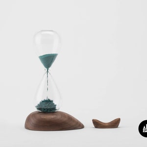 K&ouml;nnte beinhalten: Eine t&uuml;rkisfarbene Sanduhr mit einem Holzsockel und einem separaten Holzst&auml;nder. Die Sanduhr steht auf einem wei&szlig;en Hintergrund. Der Markenname "Zakkr" ist in der unteren rechten Ecke des Bildes sichtbar.