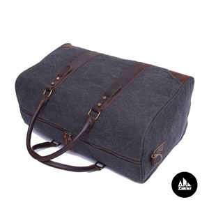 Pu&ograve; includere: Una borsa da viaggio in tela grigia con dettagli e manici in pelle marrone. La borsa ha una chiusura a zip e un anello metallico sul lato.