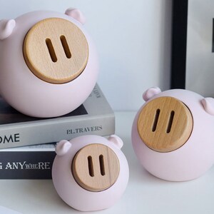 May include: Three pink ceramic piggy banks with wooden coin slots. The piggy banks are different sizes and are arranged in a row on a white surface.