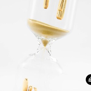 May include: A gold hourglass with a clear glass body and a gold top. The hourglass is filled with sand and is tilted on its side.