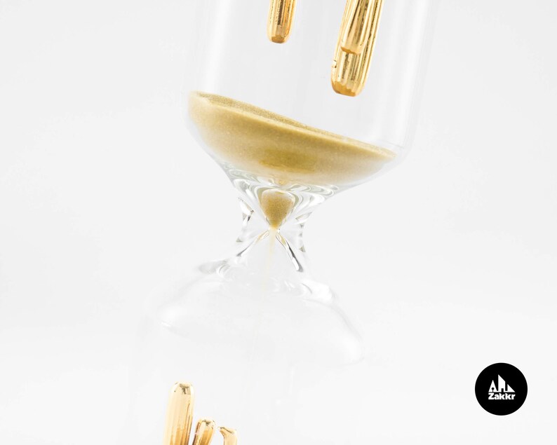 May include: A gold hourglass with a clear glass body and a gold top. The hourglass is filled with sand and is tilted on its side.