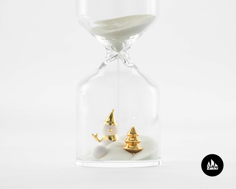 May include: A clear glass hourglass with white sand and gold-colored figurines of a snowman and a Christmas tree inside. The hourglass is on a white background.