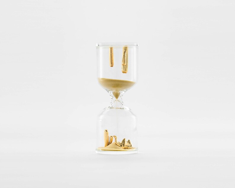 May include: A clear glass hourglass with gold accents. The top of the hourglass is filled with sand and the bottom is empty. The sand is slowly dripping from the top to the bottom.