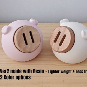 May include: Two piggy banks made of resin, one pink and one white. Each piggy bank has a wooden snout with two slots.