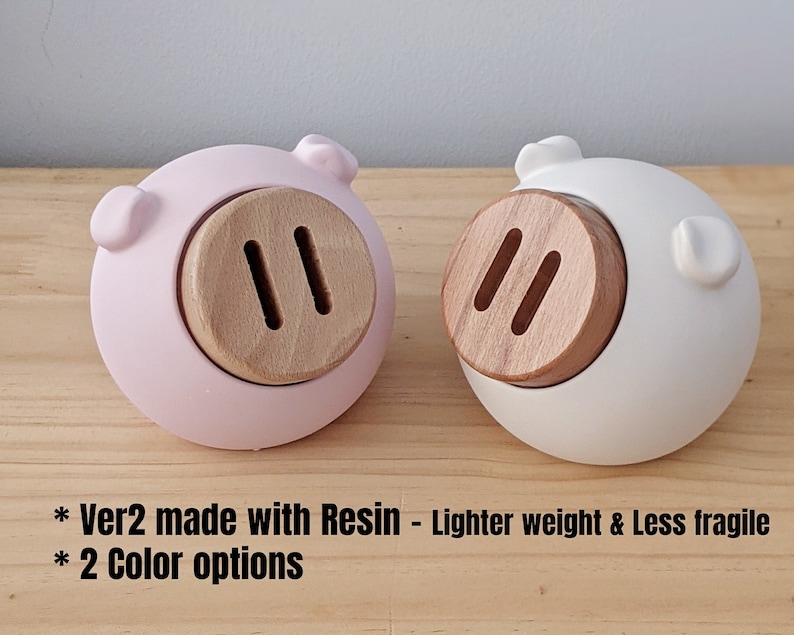 May include: Two piggy banks made of resin, one pink and one white. Each piggy bank has a wooden snout with two slots.