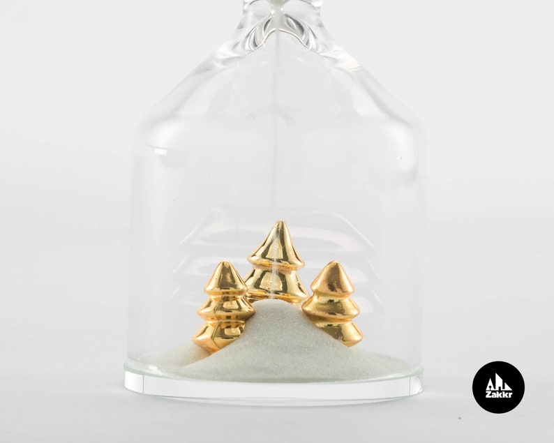 May include: A glass hourglass with a white sand base and three gold-colored trees inside. The hourglass is clear and has a rounded shape.