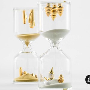May include: Two glass hourglass sand timers with gold accents. The top of the hourglass on the left has gold geometric shapes. The top of the hourglass on the right has three gold Christmas tree ornaments. The bottom of the hourglass on the left has gold geometric shapes in the sand. The bottom of the hourglass on the right has a gold snowman and a gold Christmas tree in the sand.