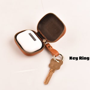 May include: A brown leather case with a zipper closure, open to reveal a white AirPod case. A key ring with a key is attached to the case.