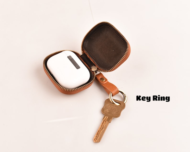 May include: A brown leather case with a zipper closure, open to reveal a white AirPod case. A key ring with a key is attached to the case.