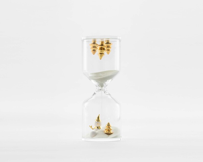 May include: A clear glass hourglass with white sand and gold-colored ornaments inside. The hourglass is on a white background.