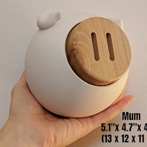 May include: White ceramic piggy bank with a wooden lid and two slots. The piggy bank measures 5.1 inches by 4.7 inches by 4.3 inches, or 13 cm by 12 cm by 11 cm.