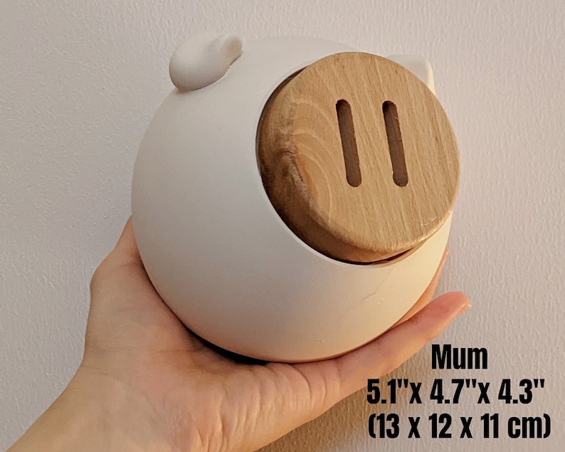 May include: White ceramic piggy bank with a wooden lid and two slots. The piggy bank measures 5.1 inches by 4.7 inches by 4.3 inches, or 13 cm by 12 cm by 11 cm.