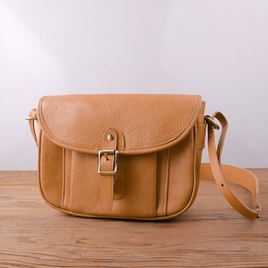 Può includere: Una borsa a tracolla in pelle color cuoio con chiusura a fibbia e una lunga tracolla regolabile. La borsa è appoggiata su una superficie di legno.