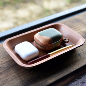 May include: A brown leather tray with a white wireless charging case, a brown and green leather case with a zipper, and a gold pen.
