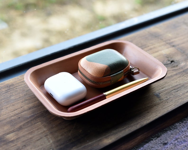 May include: A brown leather tray with a white wireless charging case, a brown and green leather case with a zipper, and a gold pen.