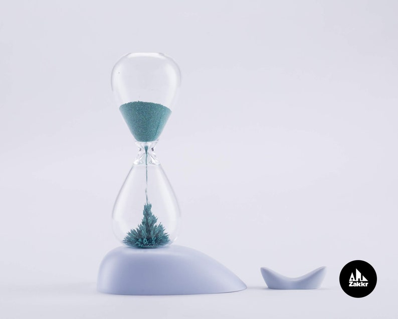 K&ouml;nnte beinhalten: Eine blaue Sanduhr mit einem blauen Sandtimer im Inneren, die auf einer wei&szlig;en Walfigur sitzt. Die Sanduhr steht auf einer wei&szlig;en Oberfl&auml;che. Die Walfigur hat eine kleine Schwanzflosse. Der Text "Zakkr" befindet sich auf einem schwarzen Kreis in der unteren rechten Ecke des Bildes.