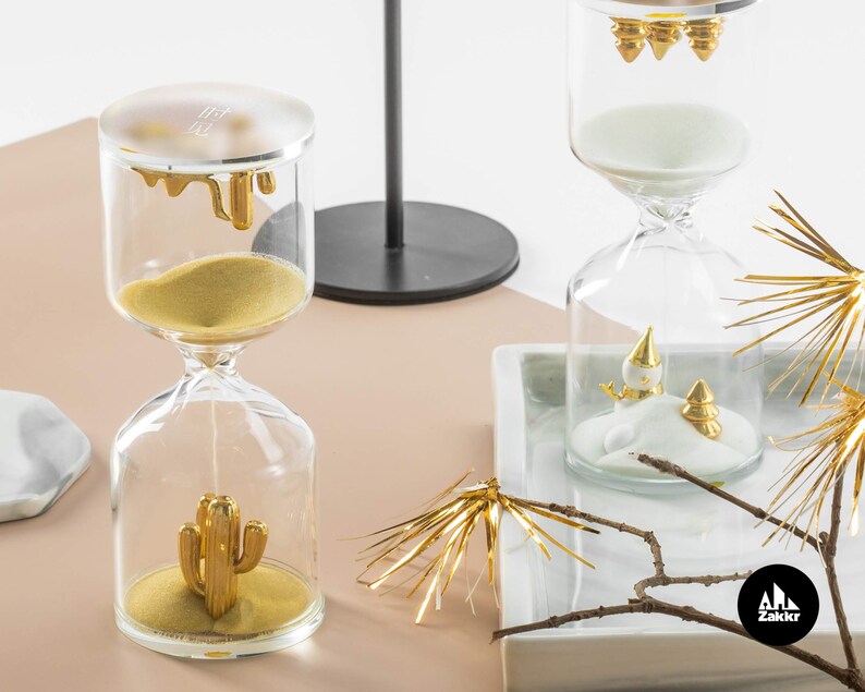 May include: Two glass hourglass sand timers with gold accents. One hourglass has a gold cactus design and the other has a gold snowman design.