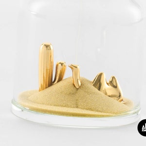 May include: A gold-colored camel, cactus, and sand dune figurine inside a clear glass dome. The figurine is a decorative piece for a home or office.