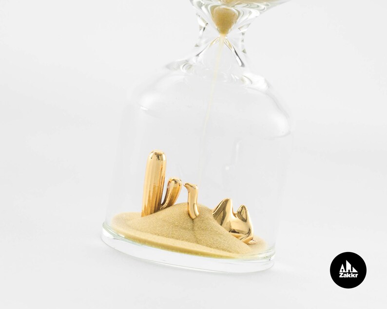 May include: A glass hourglass with gold sand and a miniature desert scene inside. The scene includes a cactus and a camel, all made of gold.