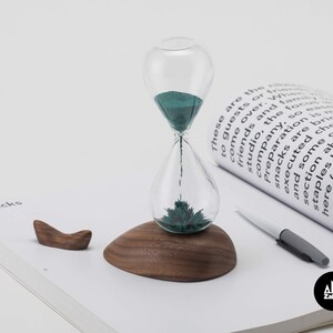K&ouml;nnte beinhalten: Eine Sanduhr mit gr&uuml;nem Sand und einem Holzsockel steht auf einer wei&szlig;en Seite mit dem aufgedruckten Wort "Snacks". Die Sanduhr besteht aus Glas und hat einen Metallsockel. Der Sand ist dunkelgr&uuml;n. Der Holzsockel ist dunkelbraun. Die Seite ist wei&szlig; und hat einen hellgrauen Hintergrund.