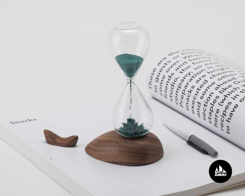 K&ouml;nnte beinhalten: Eine Sanduhr mit gr&uuml;nem Sand und einem Holzsockel steht auf einer wei&szlig;en Seite mit dem aufgedruckten Wort "Snacks". Die Sanduhr besteht aus Glas und hat einen Metallsockel. Der Sand ist dunkelgr&uuml;n. Der Holzsockel ist dunkelbraun. Die Seite ist wei&szlig; und hat einen hellgrauen Hintergrund.