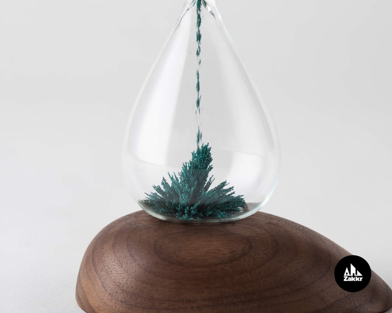 K&ouml;nnte beinhalten: Eine gl&auml;serne Sanduhr mit einem Holzsockel. Der Sand ist in einem leuchtenden T&uuml;rkisgr&uuml;n gef&auml;rbt und flie&szlig;t durch die obere Birne.