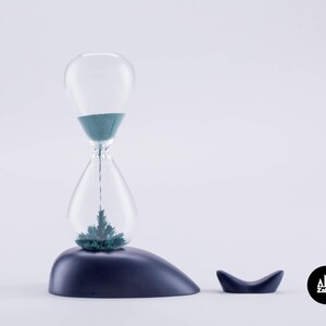 K&ouml;nnte beinhalten: Eine blaue Sanduhr mit einer klaren Glasbirne und einem dunkelblauen Sockel. Die Sanduhr ist mit gr&uuml;nem Sand gef&uuml;llt und steht auf einem dunkelblauen, walf&ouml;rmigen Sockel. Der Sockel hat einen kleinen Wal-Schwanz an der Seite. Die Sanduhr ist ein Zakkr-Produkt.