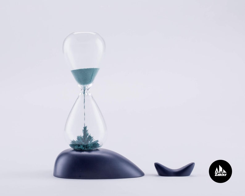 K&ouml;nnte beinhalten: Eine blaue Sanduhr mit einer klaren Glasbirne und einem dunkelblauen Sockel. Die Sanduhr ist mit gr&uuml;nem Sand gef&uuml;llt und steht auf einem dunkelblauen, walf&ouml;rmigen Sockel. Der Sockel hat einen kleinen Wal-Schwanz an der Seite. Die Sanduhr ist ein Zakkr-Produkt.