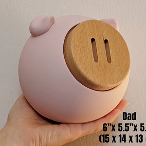 May include: A pink ceramic piggy bank with a wooden coin slot. The piggy bank is 6 inches by 5.5 inches by 5.1 inches, or 15 centimeters by 14 centimeters by 13 centimeters.